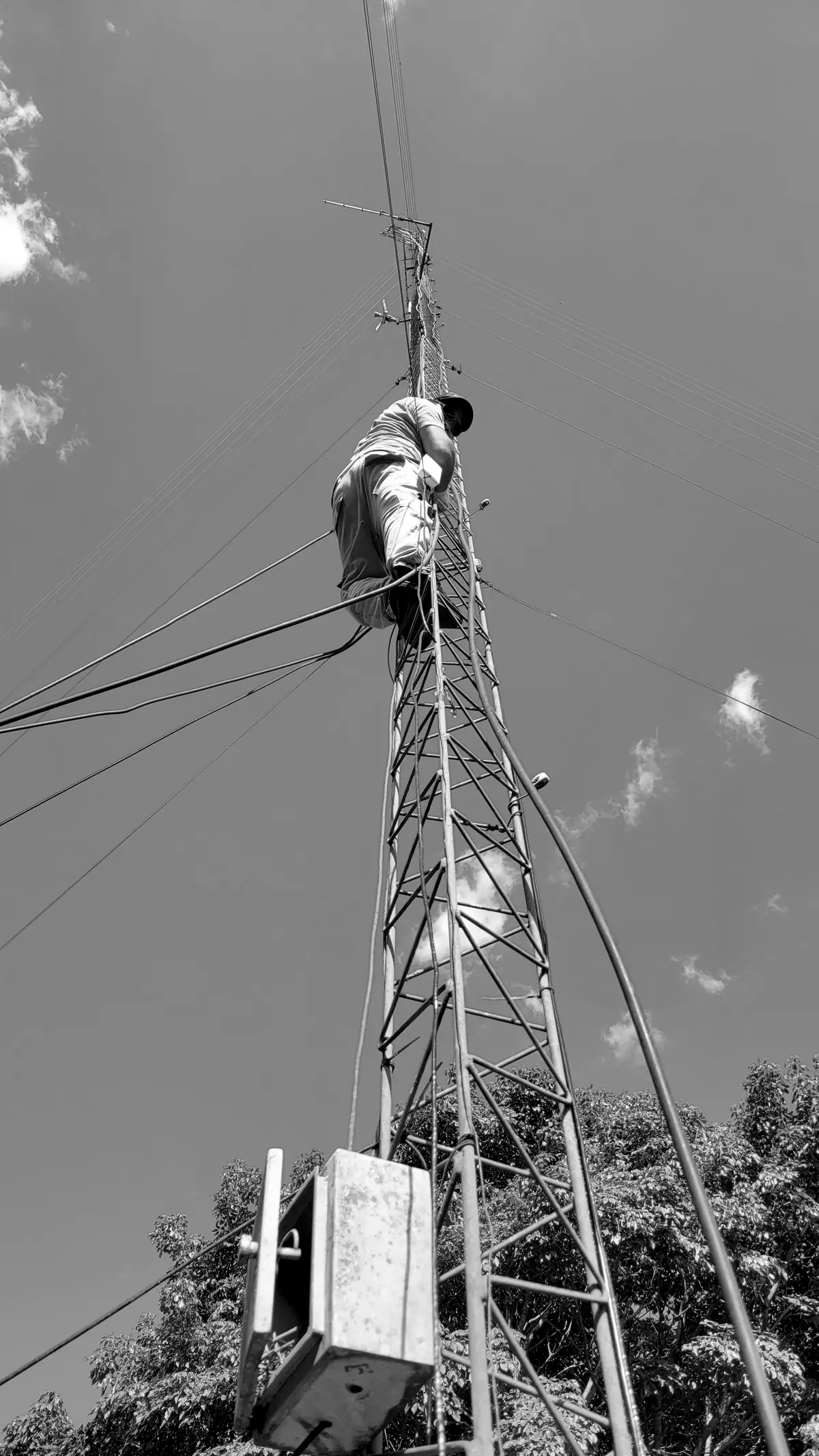 A climber at the top of a tall lattice communications tower, seen from below against the sky
