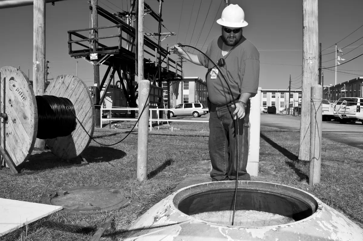 A worker in a hard hat feeding cable into an underground access point, with a large cable reel beside them