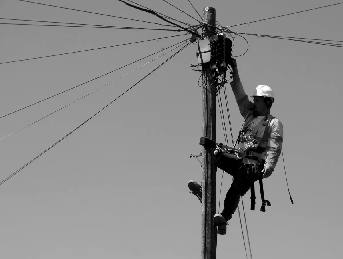 A line technician in a hard hat and safety harness working at the top of a utility pole against an open sky