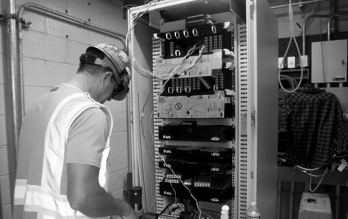 A technician in a hard hat and high-visibility vest working at an open telecommunications equipment rack